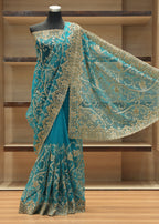 A teal blue saree with intricate gold floral embroidery and a wide matching border, displayed on a black mannequin in front of wooden shelving.