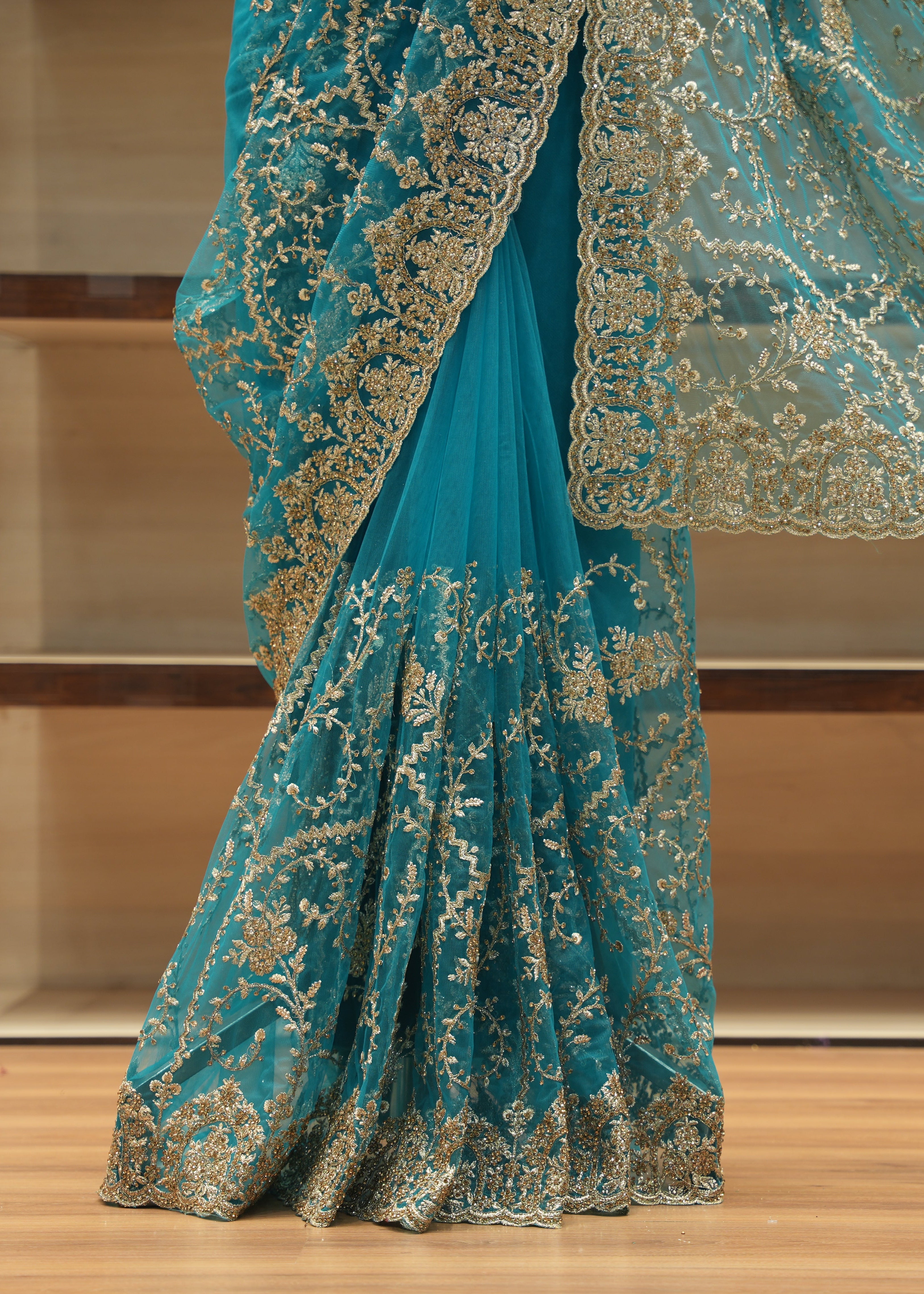 A close-up of a teal blue saree with intricate gold floral embroidery and a matching sheer dupatta draped over a wooden shelf background.