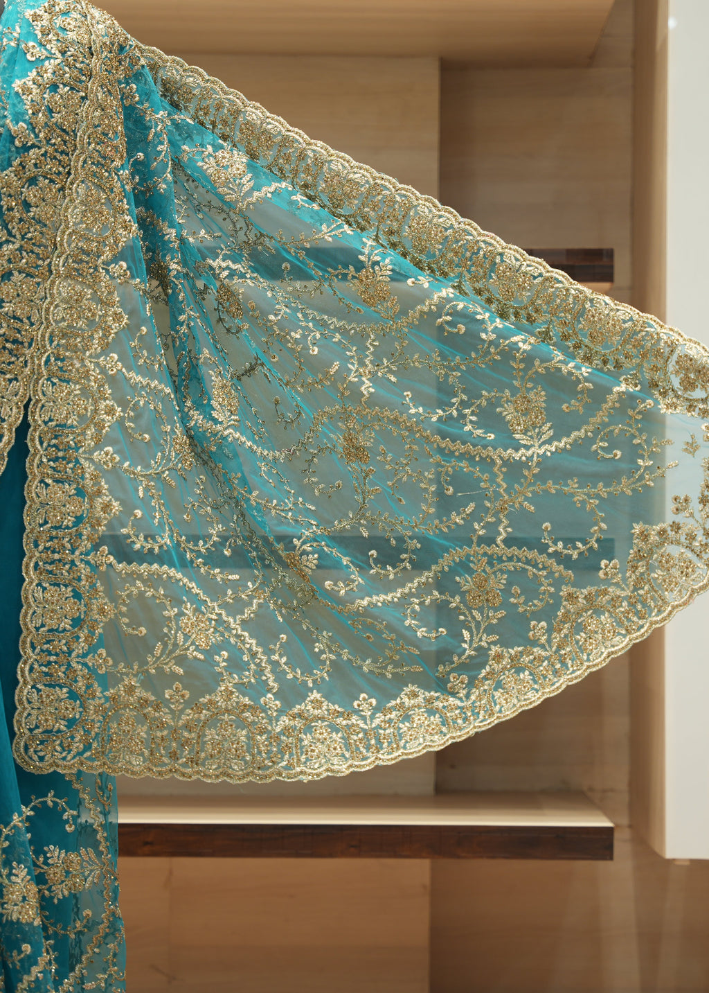 A close-up of a teal blue embroidered fabric, likely a saree drape or dupatta, featuring intricate gold floral embroidery and a scalloped border, shown against a blurred wooden background.