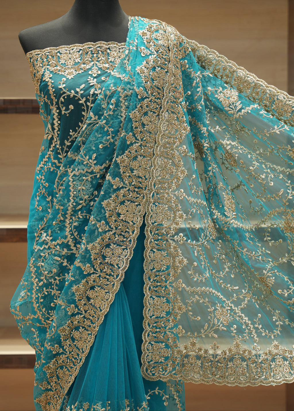 Turquoise blue saree with intricate gold floral embroidery and a matching sheer dupatta draped on a black mannequin torso against a wooden shelving background.
