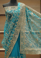 Turquoise blue saree with intricate gold floral embroidery and a matching sheer dupatta draped on a black mannequin torso against a wooden shelving background.