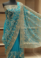 Turquoise blue saree with intricate gold floral embroidery and a matching sheer dupatta, displayed on a black mannequin torso against a wooden shelving background.