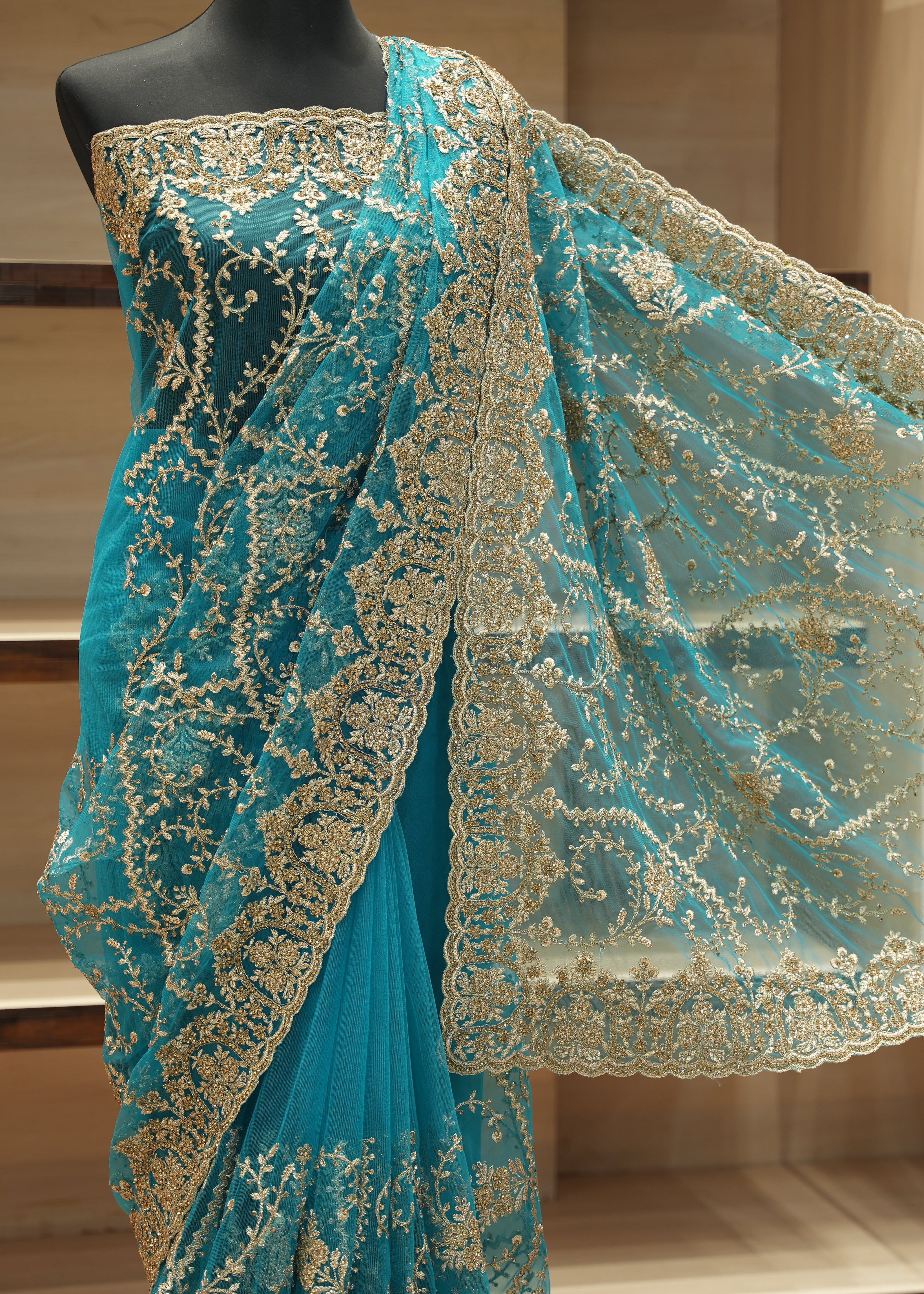Turquoise blue saree with intricate gold floral embroidery and a matching sheer dupatta, displayed on a black mannequin torso against a wooden shelving background.