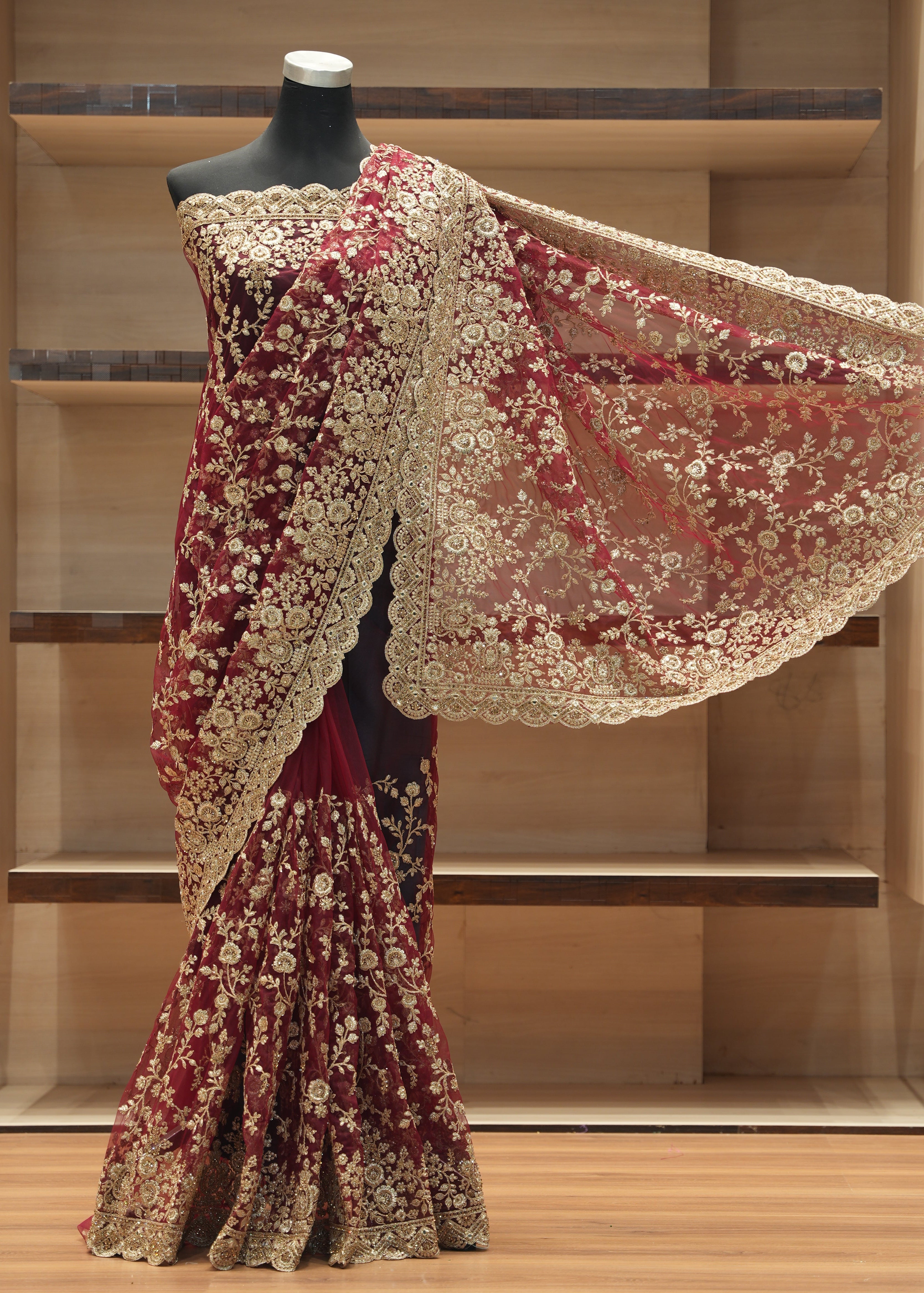 A heavily embroidered sari displayed on a black mannequin torso in front of wooden shelving. The sari is deep maroon with extensive gold floral embroidery and a wide matching gold embroidered border.