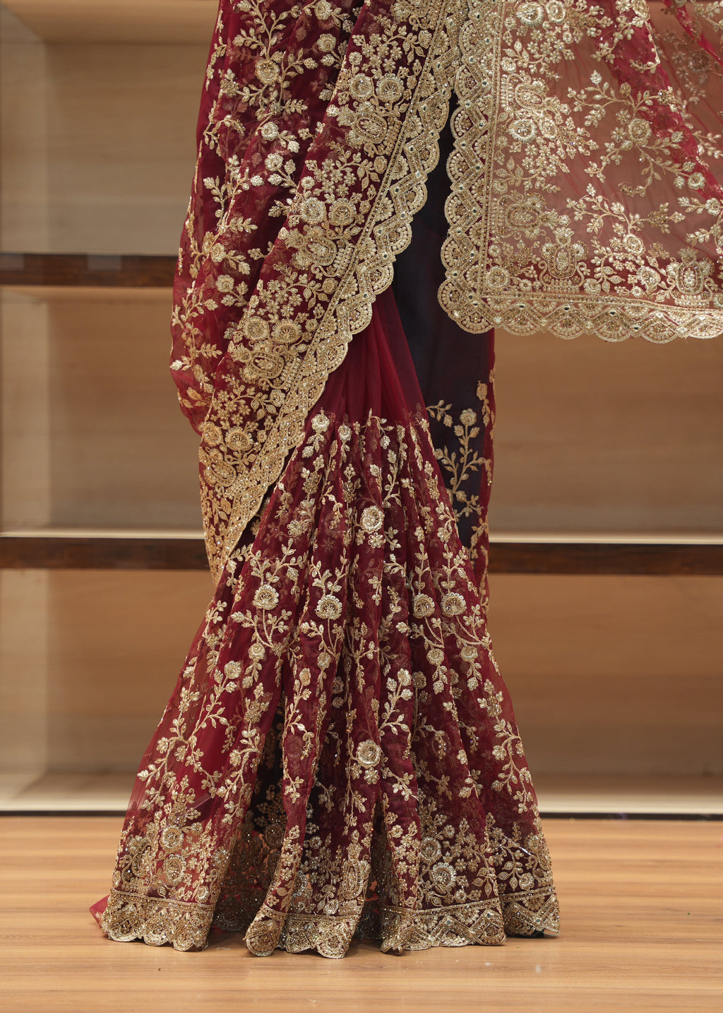 A close-up of a traditional Indian bridal lehenga skirt in deep maroon velvet, heavily embellished with intricate gold floral embroidery and a matching sheer dupatta draped over the shoulder, set against a wooden shelving background.