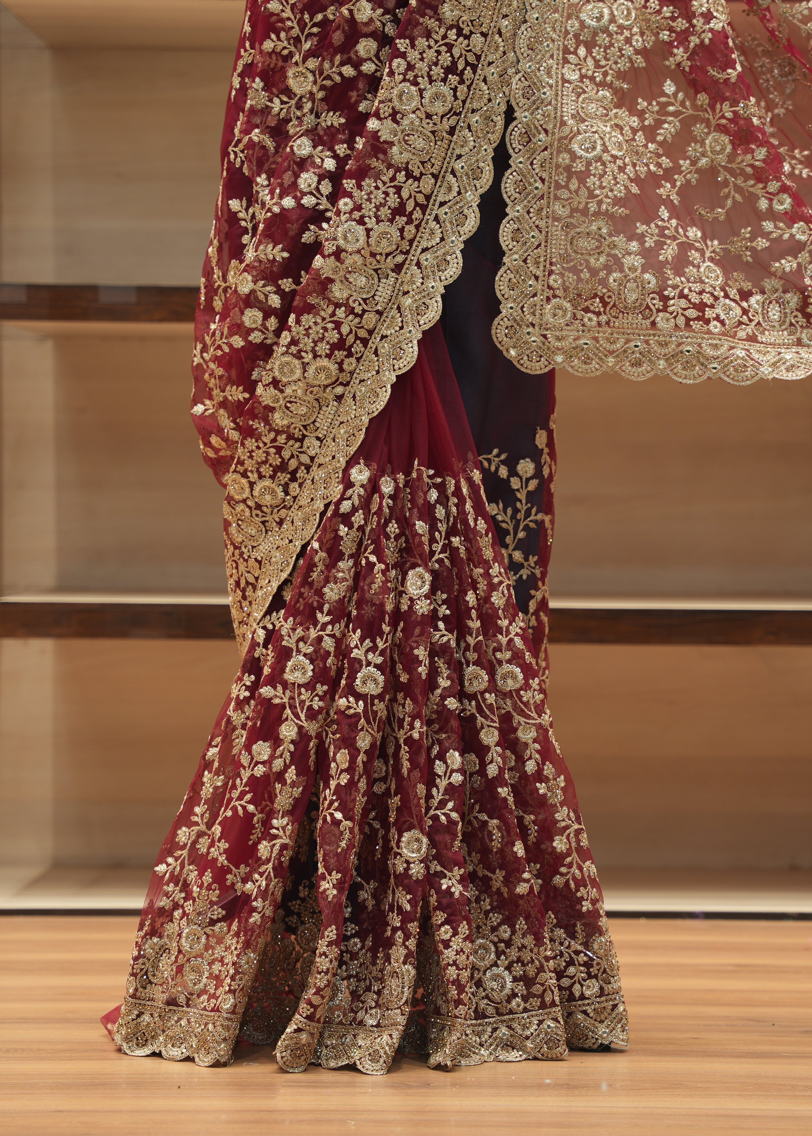 A close-up of a traditional Indian bridal lehenga skirt in deep maroon velvet, heavily embellished with intricate gold floral embroidery and a matching sheer dupatta draped over the shoulder, set against a wooden shelving background.