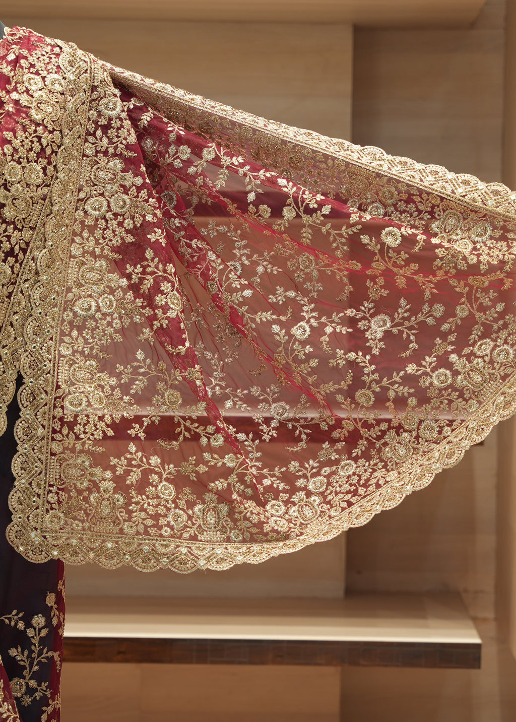 A close-up of a rich burgundy red sheer mesh fabric with extensive gold floral embroidery and a scalloped gold border, draped over a dark red embroidered garment. The fabric is displayed against a background of wooden shelving.