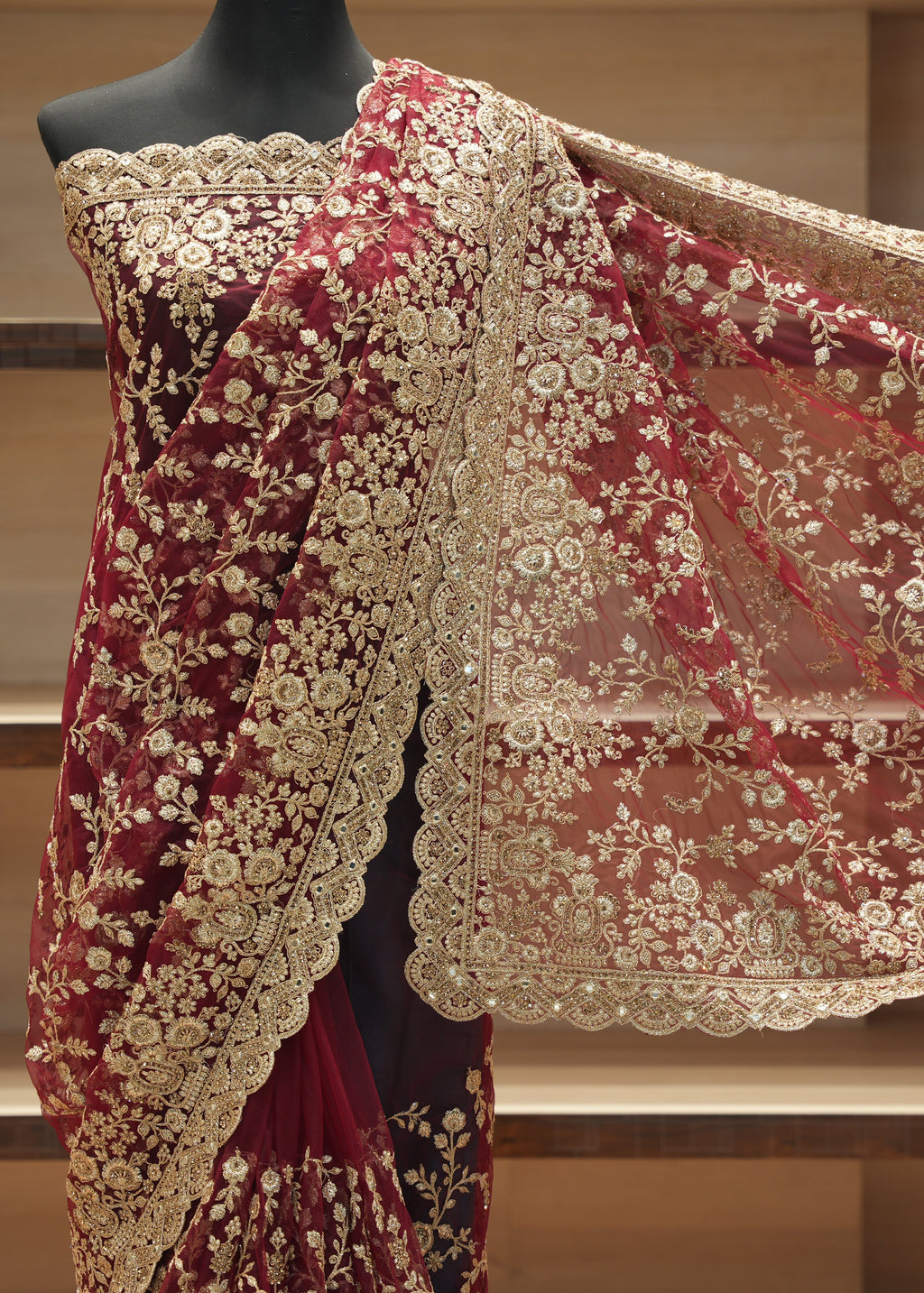 A deep maroon velvet saree with intricate gold floral embroidery and a matching sheer dupatta, displayed on a black mannequin torso against a wooden shelving background.