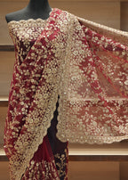 A deep maroon velvet saree with intricate gold floral embroidery and a matching sheer dupatta, displayed on a black mannequin torso against a wooden shelving background.