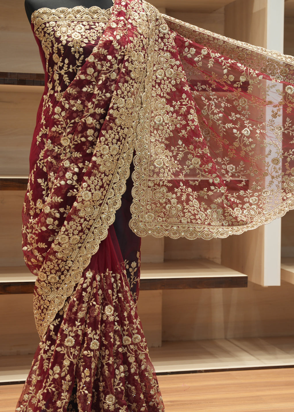 A maroon and gold embroidered saree draped on a mannequin, with the matching sheer dupatta spread out to show its intricate floral lace border, set against a wooden shelving background.