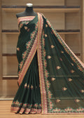 A dark forest green saree with intricate pink, gold, and white floral borders and motifs, displayed on a black mannequin torso in front of wooden shelving.
