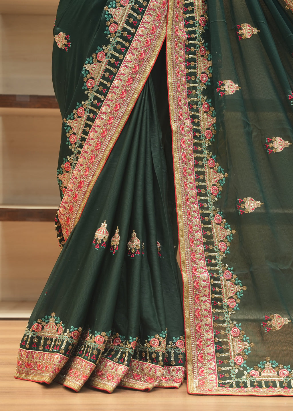 A dark forest green georgette saree with intricate floral and geometric embroidery in pink, red, and gold, draped elegantly on a wooden shelf.