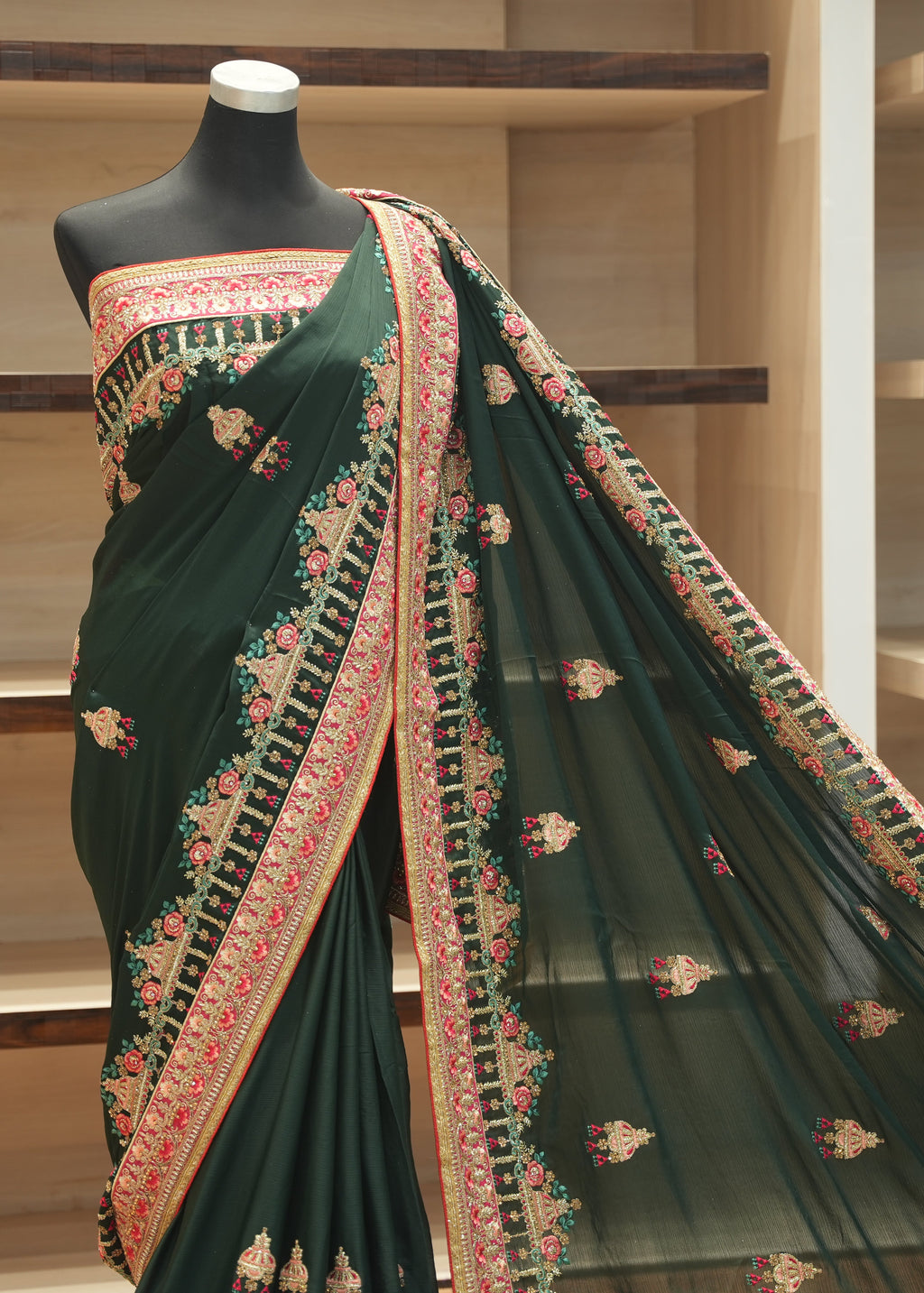 A dark forest green saree with intricate gold, pink, and green floral embroidery, displayed on a black mannequin torso in front of wooden shelves.