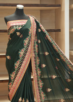 A dark forest green saree with intricate gold, pink, and green floral embroidery, displayed on a black mannequin torso in front of wooden shelves.