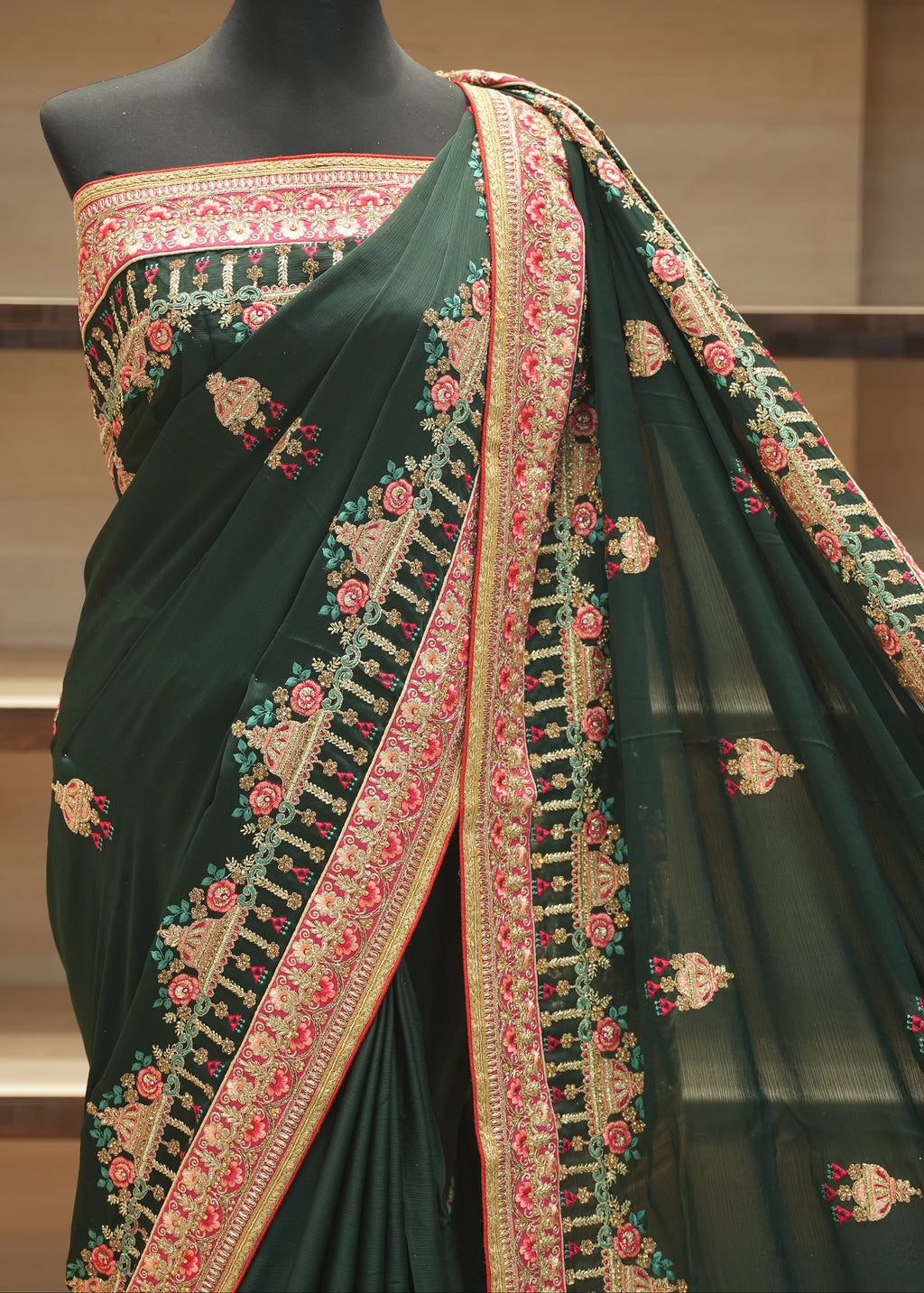 A dark forest green saree with intricate gold, pink, and red floral embroidery, displayed on a black mannequin torso against a wooden shelf background.