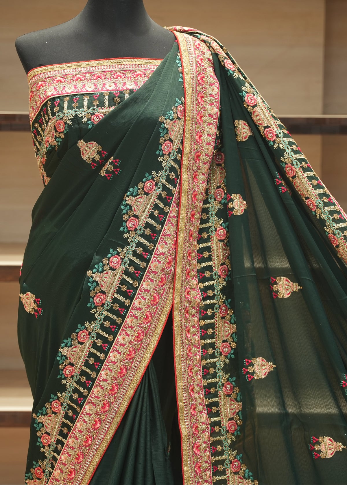 A dark forest green saree with intricate gold, pink, and red floral embroidery, displayed on a black mannequin torso against a wooden shelf background.