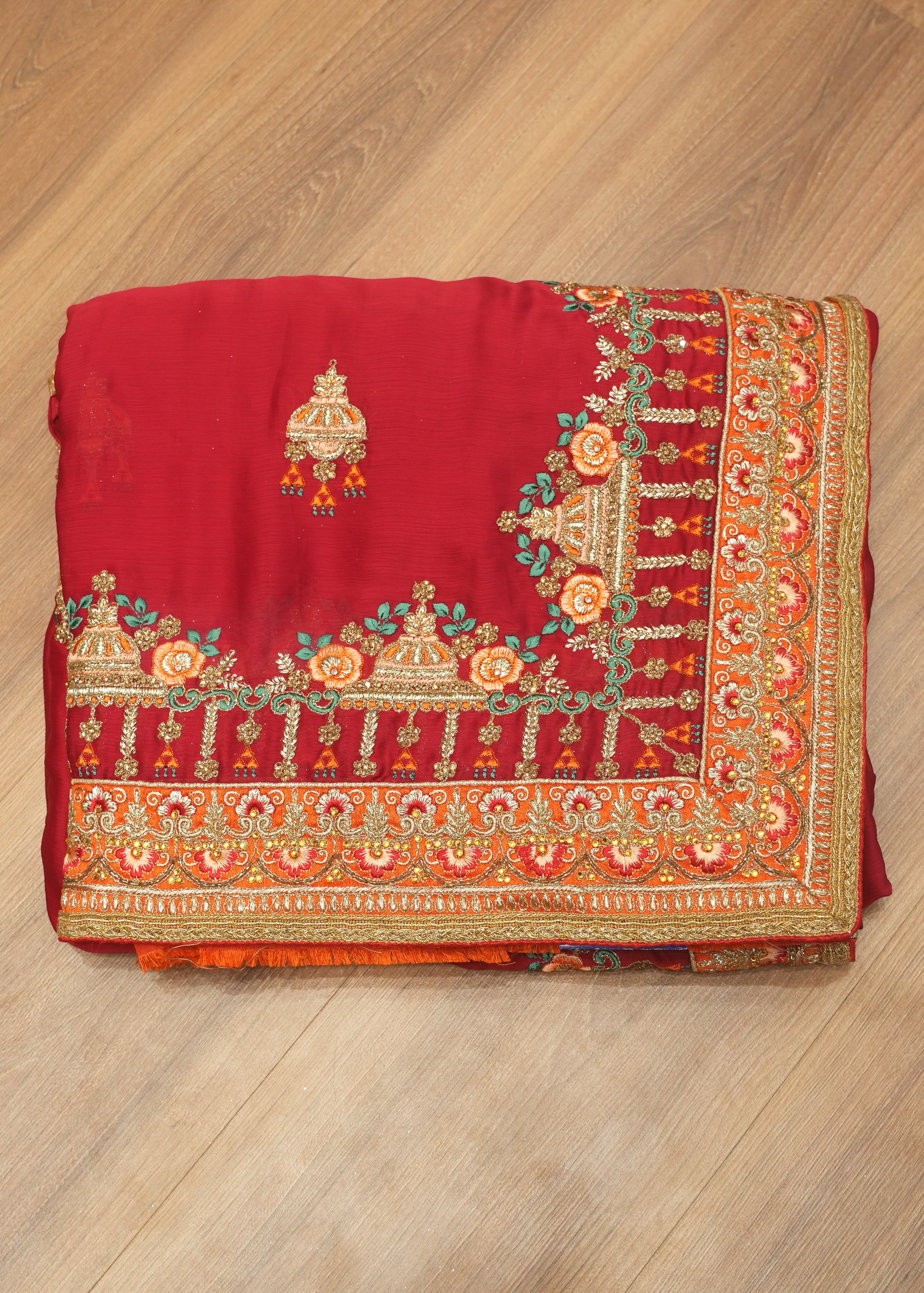 A folded red fabric piece with ornate gold, orange, and teal embroidery, displayed flat on a light brown wooden floor.