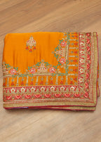 A folded orange traditional fabric with intricate gold, red, and green embroidery, displayed on a light brown wooden floor.