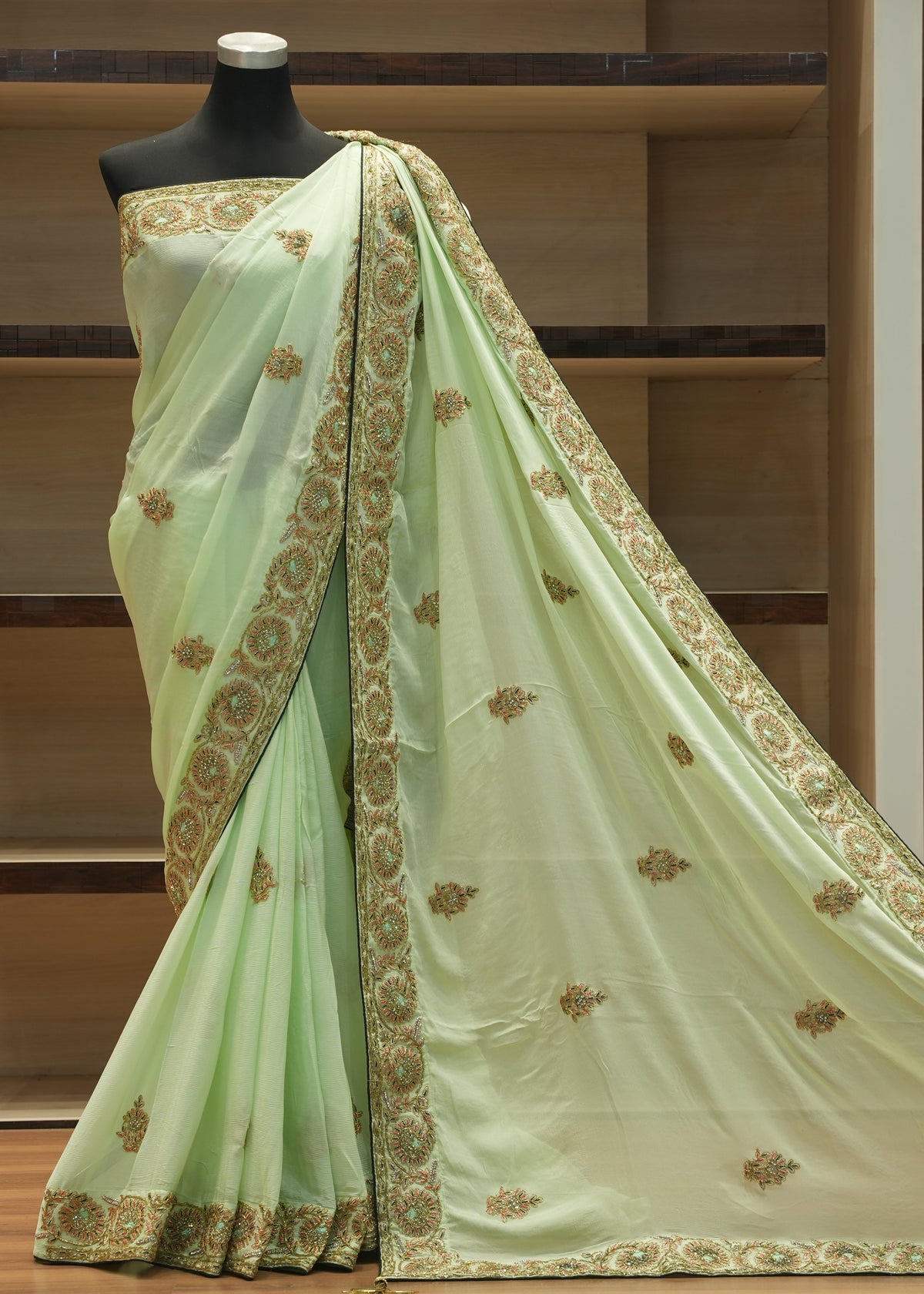A light green saree draped on a black mannequin, featuring a wide gold and green border and scattered gold floral motifs, set against a wooden shelving background.