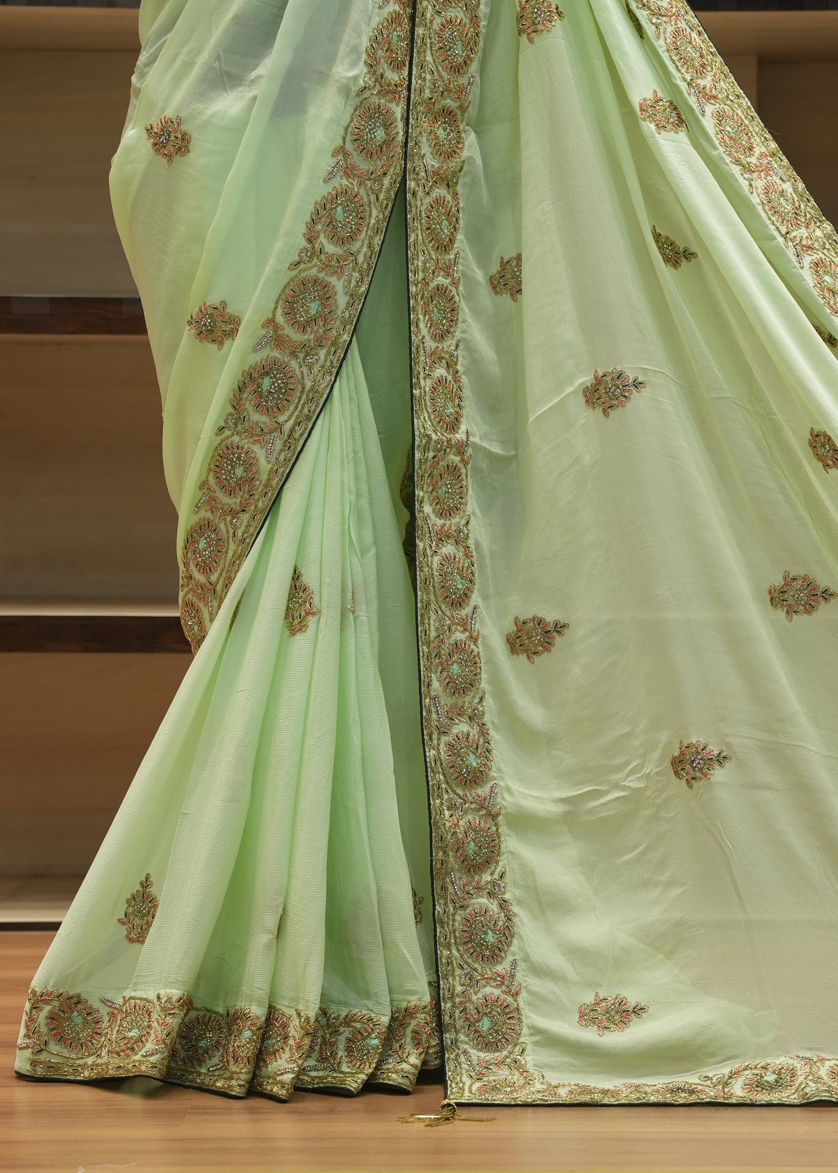 A light green saree with intricate gold and brown floral embroidery along the border and scattered motifs on the body, draped over a wooden stand.