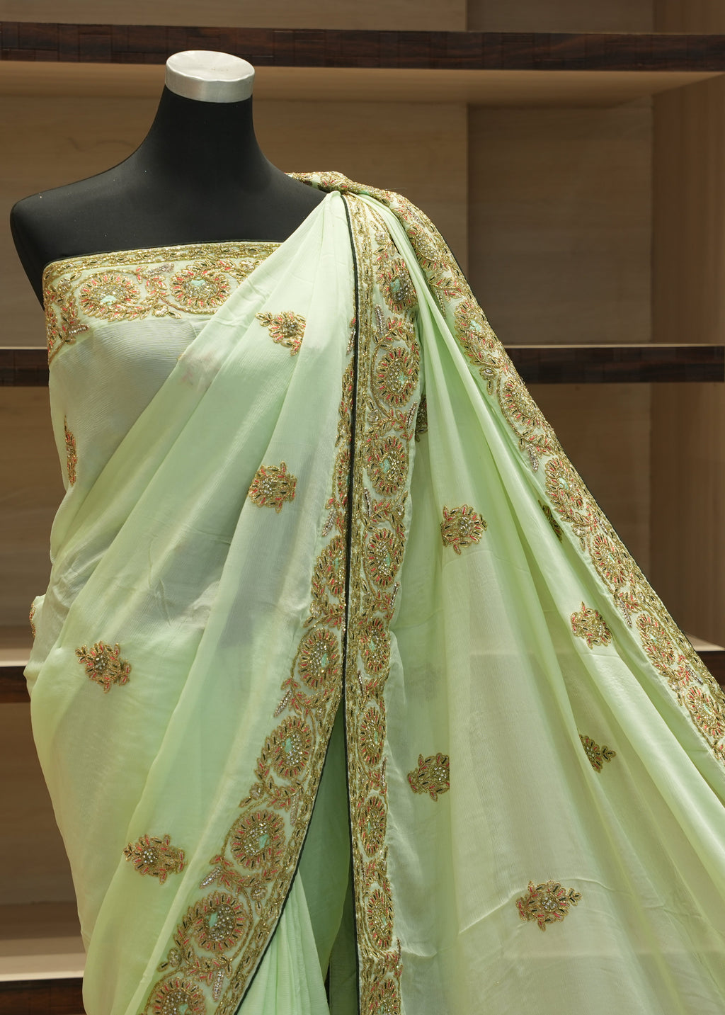 A light green saree with gold and brown floral embroidery, draped on a black mannequin torso in front of wooden shelves.