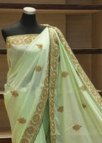 A light green saree with gold and brown floral embroidery, draped on a black mannequin torso in front of wooden shelves.