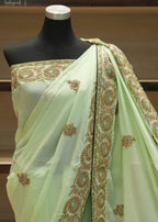 A pale mint green saree with intricate gold and green floral embroidery, draped on a black mannequin torso against a wooden shelf background.