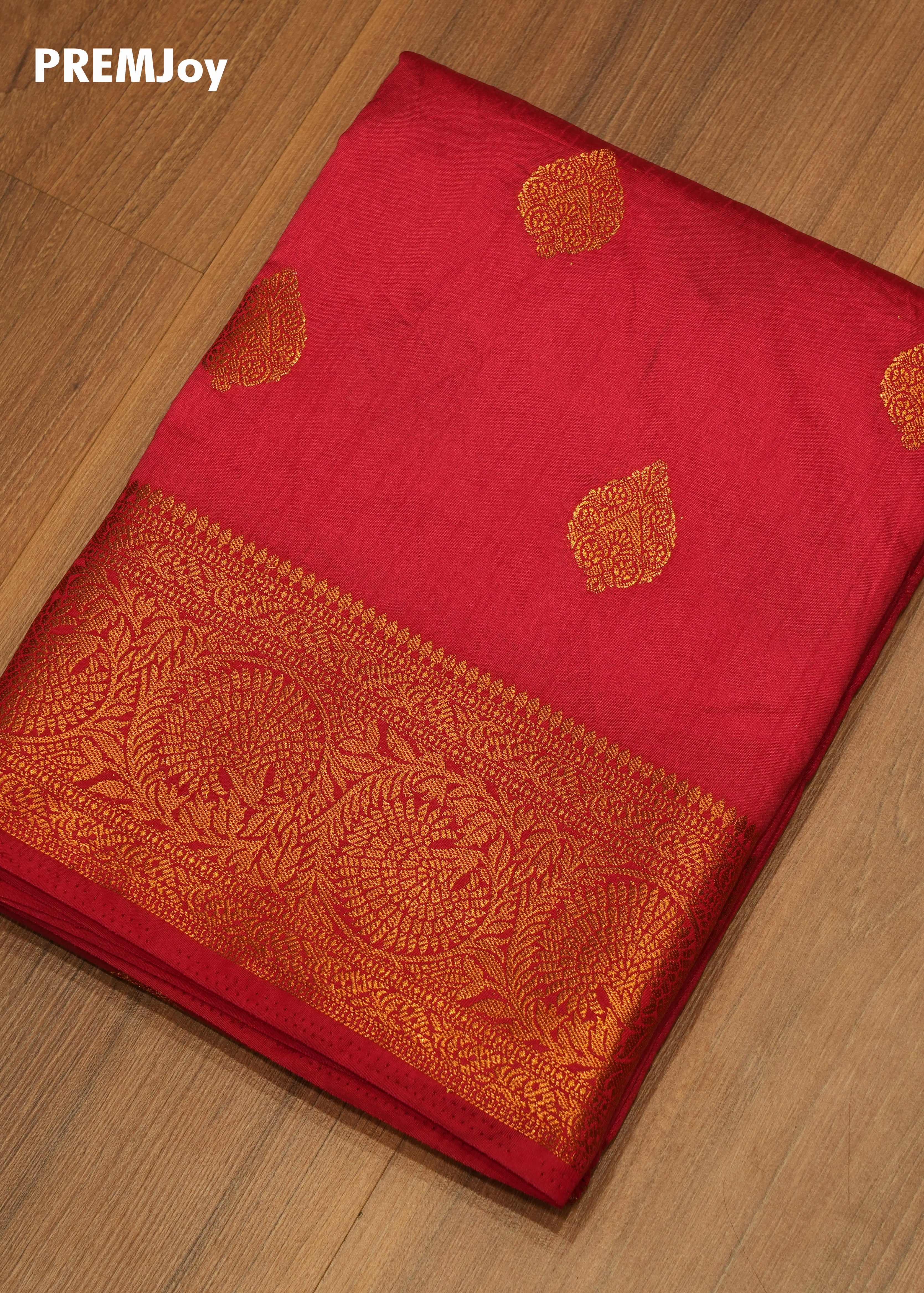 Red saree with gold embroidery on a wooden surface, branded 'PREMJoy'.