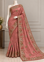 A dusty pink saree with intricate gold, green, and red floral embroidery, displayed on a white mannequin in an elegant indoor setting.