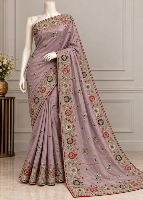 A mauve purple saree with intricate multicolor floral embroidery and a wide decorative border, displayed on a white mannequin in an elegant indoor setting.